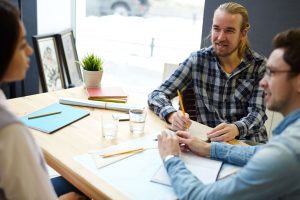 Três alunos de Engenharia em uma mesa, falando sobre um projeto. Na mesa é possível ver papéis, lápis e esquadros espalhados.