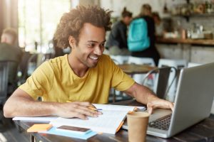 Aluno de graduação fazendo pesquisa de iniciação científica sobre uma mesa. Há papéis espalhados e ele usa um notebook.