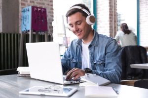 Estudante cria sua apresentação em seu notebook, na mesa, podemos ver diversos papéis e uma caneta.